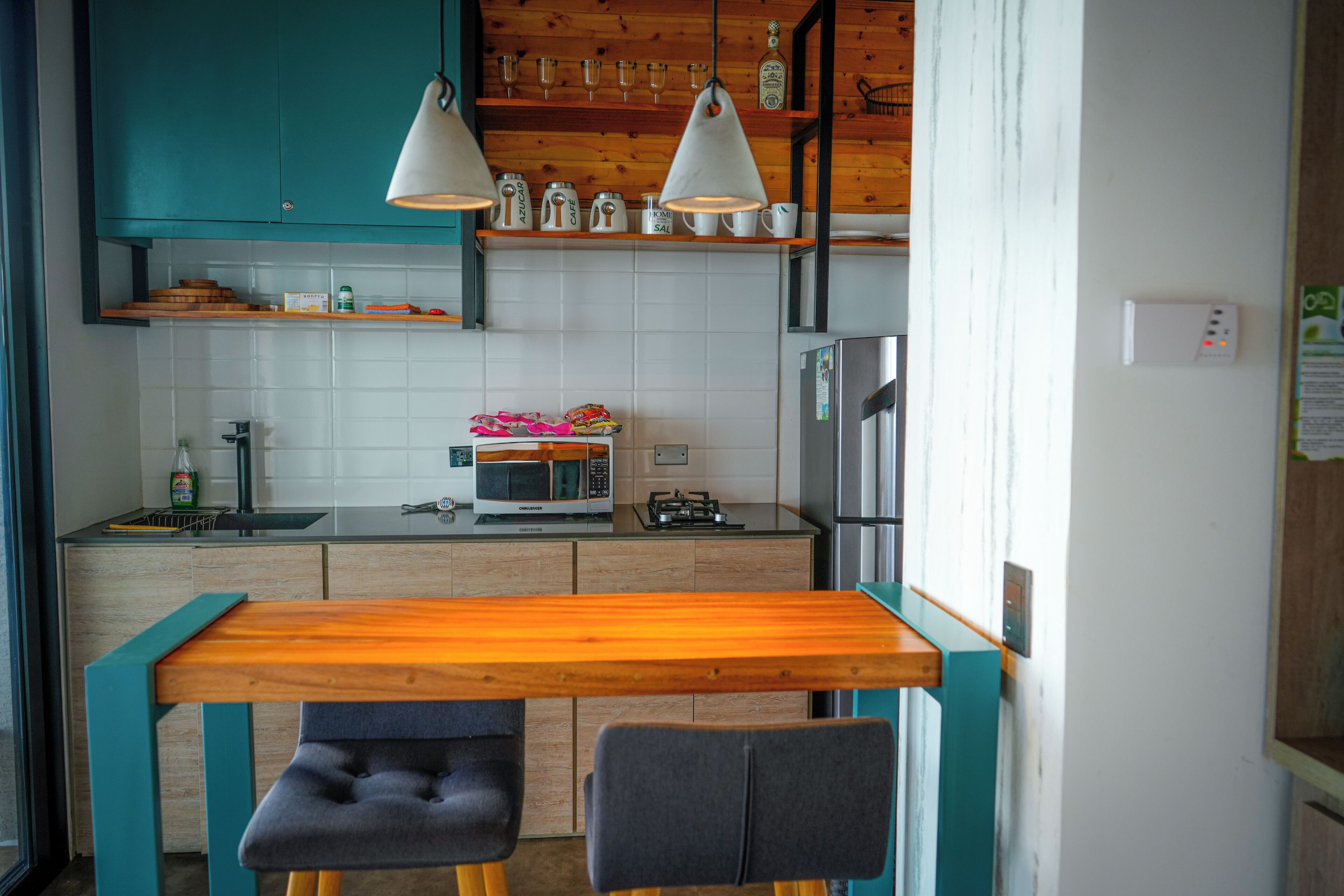 Una cocina compacta con armarios color turquesa, una mesa de madera, dos sillas, un microondas, un refrigerador y luces colgantes.