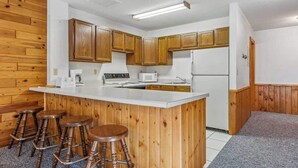A kitchen with wooden cabinets, a white countertop, a microwave, a stove, and three stools. - Lakeside Condo #17 3BR 3BA Timberlane Resort (Park Rapids)