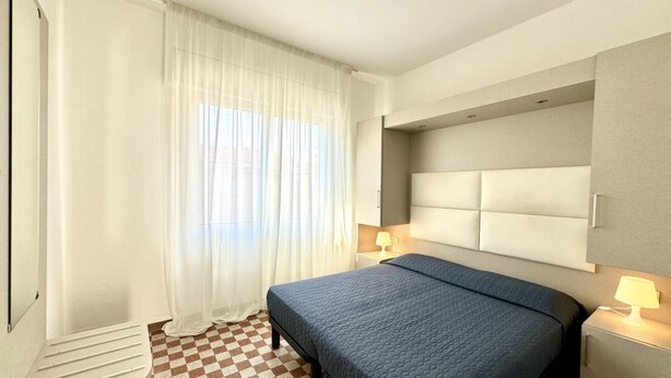 A hotel room with a bed, a nightstand, a lamp, a window with curtains, and a checkered floor. - Villa Bianca 6 - Carraro Immobiliare Jesolo (Lido di Jesolo)