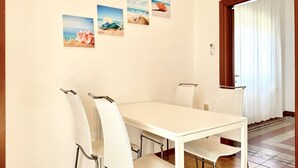 A dining area with a white table and chairs, a window with white curtains, and three beach-themed pictures on the wall.