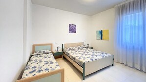 A bedroom with two single beds, a nightstand with a lamp, a small painting on the wall, and a window with sheer curtains.