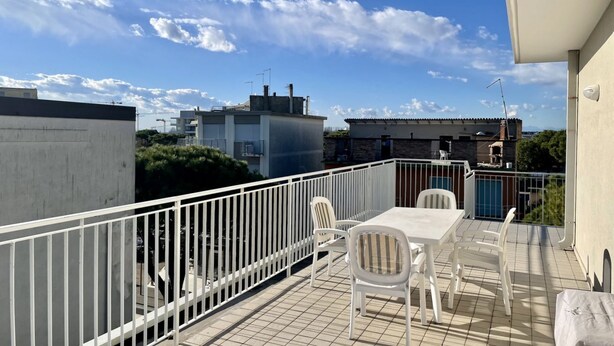 A rooftop terrace with a table and chairs, a white railing, and a view of surrounding buildings and trees.