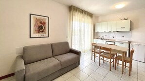 A living room with a sofa, a dining table with chairs, a kitchen with cabinets, and a framed picture on the wall.
