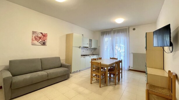 Una sala de estar con sofá, una mesa de comedor con sillas, una zona de cocina y un televisor montado en la pared.