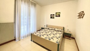 Un dormitorio con cama, mesita de noche, radiador y ventana con cortinas.