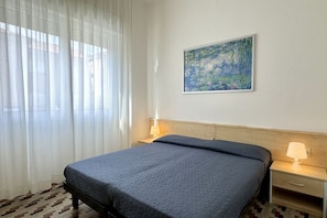 Une chambre d’hôtel avec un lit, une table de chevet, une lampe, un tableau au mur et des rideaux légers.