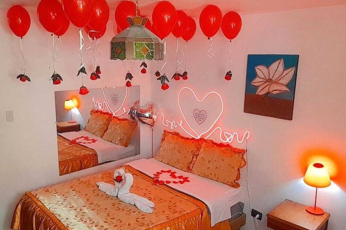 A hotel room with two beds, red balloons, a heart-shaped neon sign, and a flower painting.
