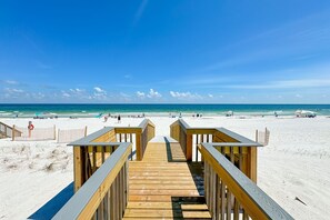 Ein hölzerner Steg führt zu einem Strand mit blauem Himmel und ruhigem Wasser.