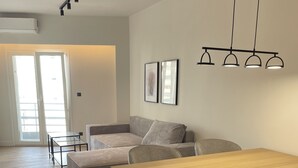 A modern living room with a wooden dining table, grey upholstered sofa, and a glass coffee table. There are framed pictures on the wall and a ceiling light fixture. - Athens City Chic apartment  in Petralona,with 2 double bedrooms and 2 bathrooms (Athens)