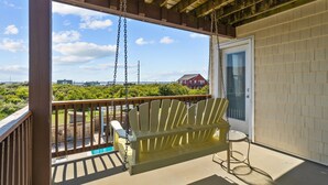 A wooden deck with a swing chair and a small table, overlooking a lush green landscape and a distant red building. - 5711-OBX TA SEA - Semi-Oceanfront Escape w/ Pool & Hot Tub (Nags Head)