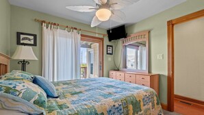 Une chambre à coucher comprenant un lit, un ventilateur de plafond, une commode, un miroir et une fenêtre avec des rideaux.