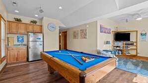 Une table de billard dans un salon, avec un coin cuisine et un téléviseur en arrière-plan.