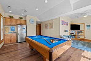 Une table de billard dans un salon, avec un coin cuisine et un téléviseur en arrière-plan.