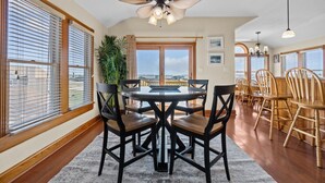 A dining area with a round table, black chairs, and a ceiling fan. There are large windows with blinds, a potted plant, and a view of the ocean. - 5711-OBX TA SEA - Semi-Oceanfront Escape w/ Pool & Hot Tub (Nags Head)
