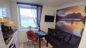 A compact living space with a kitchenette, a dining area with a table and chairs, a black leather sofa with decorative pillows, a large window with a view of the outdoors, and a mounted flat-screen TV.