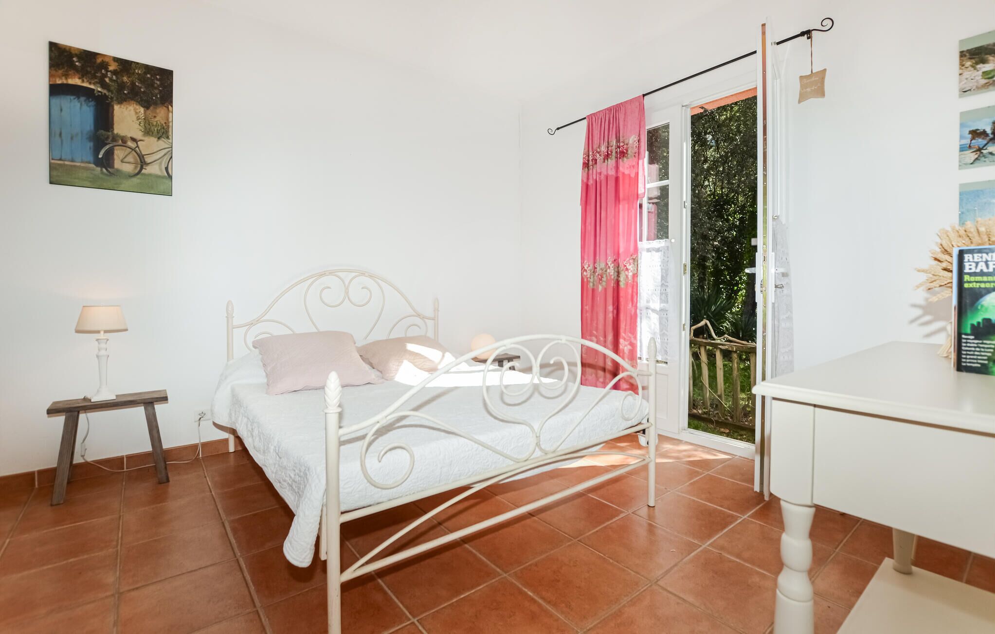 Ein Schlafzimmer mit einem weißen Metallbett, einem Nachttisch mit Lampe, einem Fenster mit rosa Vorhängen und einem Bild an der Wand.