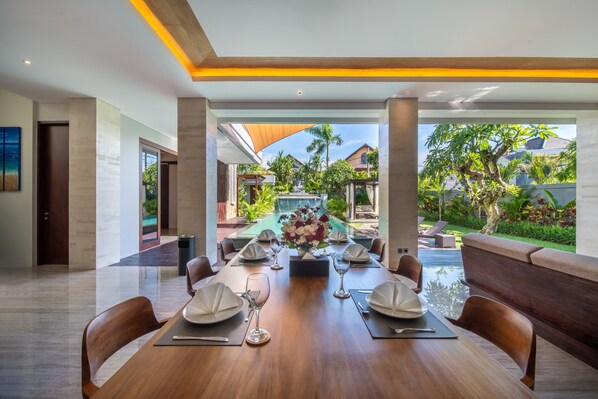 A dining area with a long table set for six, a pool in the background, and a view of a tropical garden.