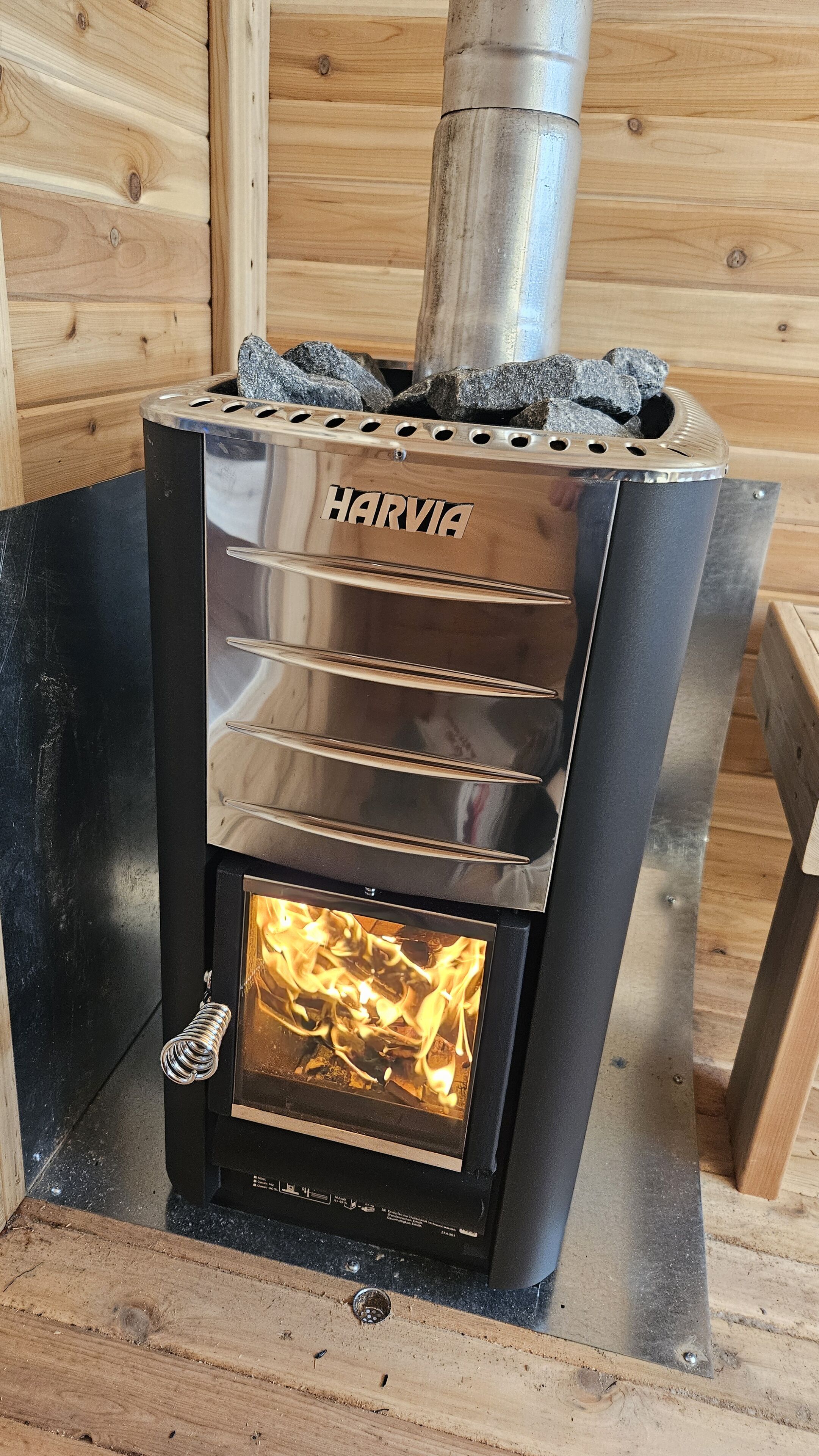 Een Harvia houtkachel met glazen voorpaneel, metalen ventilatieopeningen en schoorsteen.