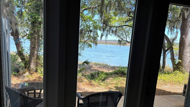 Vue depuis une pièce dotée d’une grande fenêtre donnant sur un lac, entouré d’arbres et agrémentée d’une terrasse avec des chaises et une table.