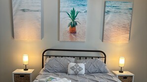 Un lit bien fait, recouvert d’une couverture grise, avec deux lampes de chevet et trois œuvres d’art mural représentant des scènes de plage.