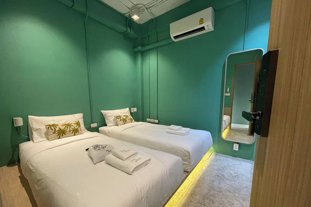 Een hotelkamer met twee bedden, elk voorzien van witte lakens en palmboomkussens. Er is een spiegel aan de muur, een airconditioner boven de bedden, en een houten deur.