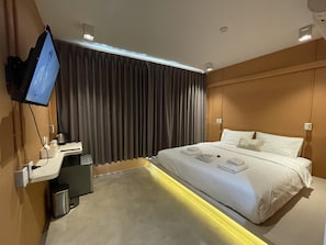 A modern hotel room with a large bed, a desk with a computer, a TV mounted on the wall, and a window with curtains.
