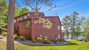 A wooden cabin with a large deck, a view of the water, and a well-maintained lawn. - Lakeside Condo #14 3BR 3BA Timberlane Resort (Park Rapids)