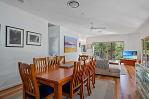 A dining area with a wooden table and chairs, a sofa, and a TV. - Large family home with a wrap around deck catching the sea breeze. (Noosa)