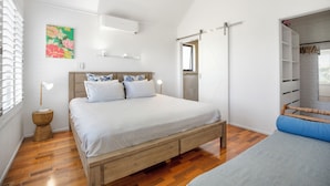 Un dormitorio con cama de madera, una mesita de noche, un armario con ropa colgada y aire acondicionado de pared. Incluye una ventana con persianas.