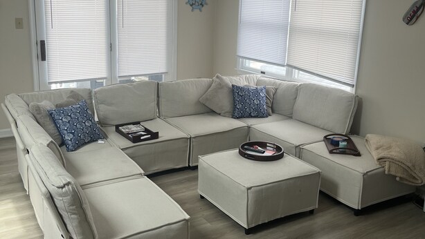 Ein modernes Wohnzimmer mit einer Eckcouch, Poufs und einem Couchtisch. Zwei große Fenster mit Jalousien, eine wandhängende Uhr und ein kleines dekoratives Accessoire sind ebenfalls vorhanden.