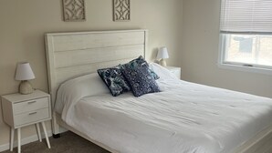 Ein Schlafzimmer mit einem weißen Bett, zwei Nachttischen, einem Fenster mit Jalousien und zwei Wandpaneelen zur Dekoration.