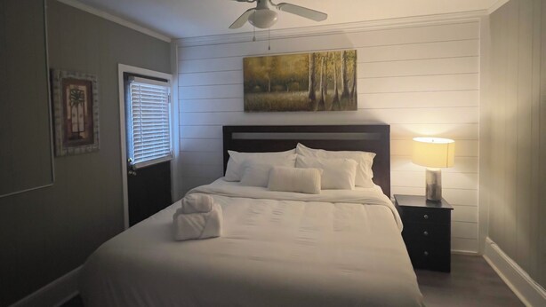 Een net opgemaakt bed in een kamer met een plafondventilator, een schilderij aan de muur en een nachtkastje met een lamp.