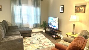 Een woonkamer met een grijze bank, een bruine fauteuil, een televisie op een houten dressoir, een vloerkleed met dessin en een lamp op een bijzettafeltje.