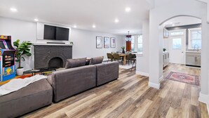 Uma sala de estar moderna com um sofá seccional cinza, uma televisão de tela plana, uma lareira e uma área de jantar com mesa e cadeiras.