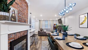 Cozinha moderna com lareira de tijolos, sala de jantar com mesa de madeira posta para quatro e sala de estar com sofá e uma planta grande.