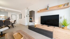 A modern living room with a flat-screen TV, wooden furniture, and a dining area in the background.
