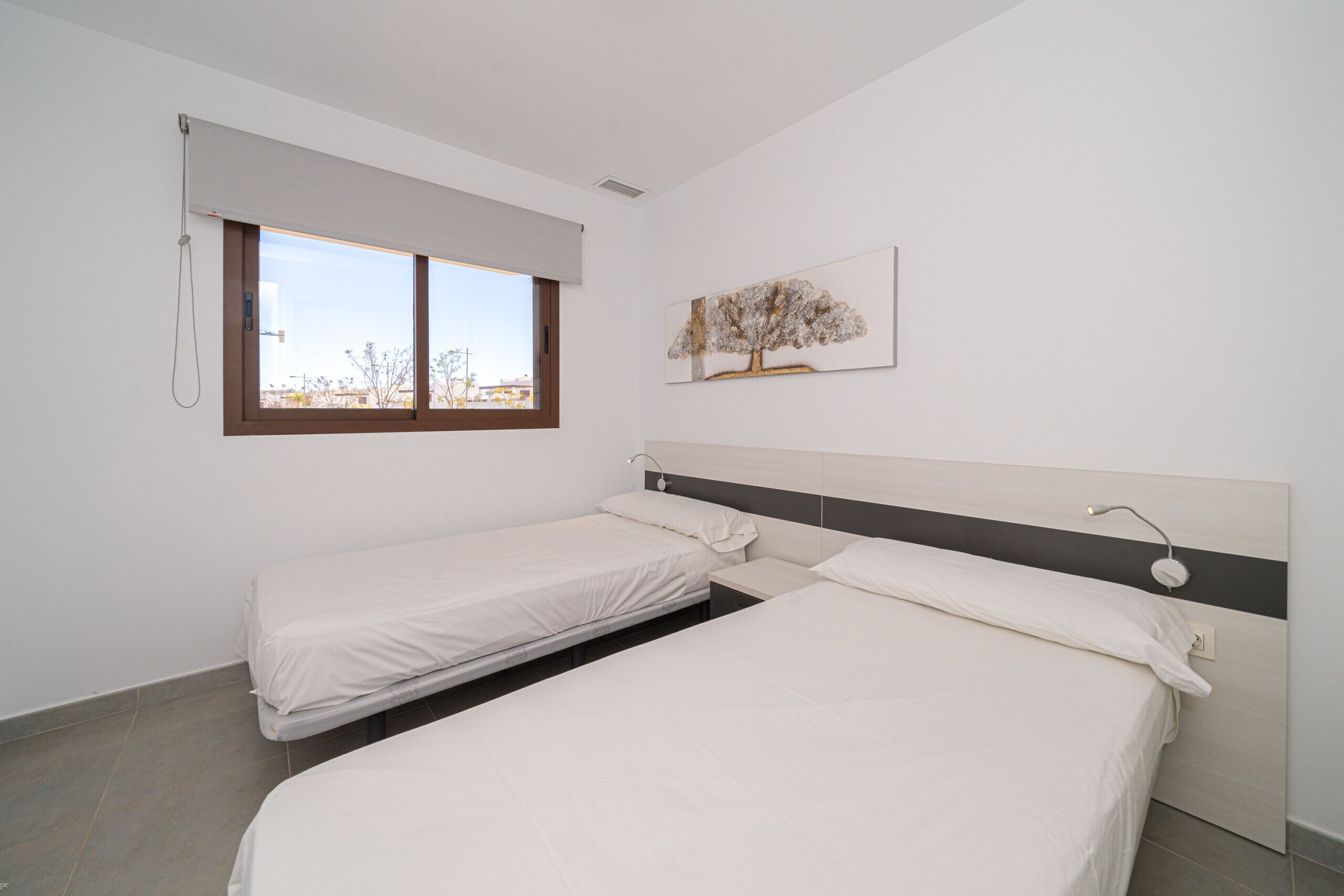 A hotel room with two single beds, a window with a view, a painting on the wall, and a ceiling light.