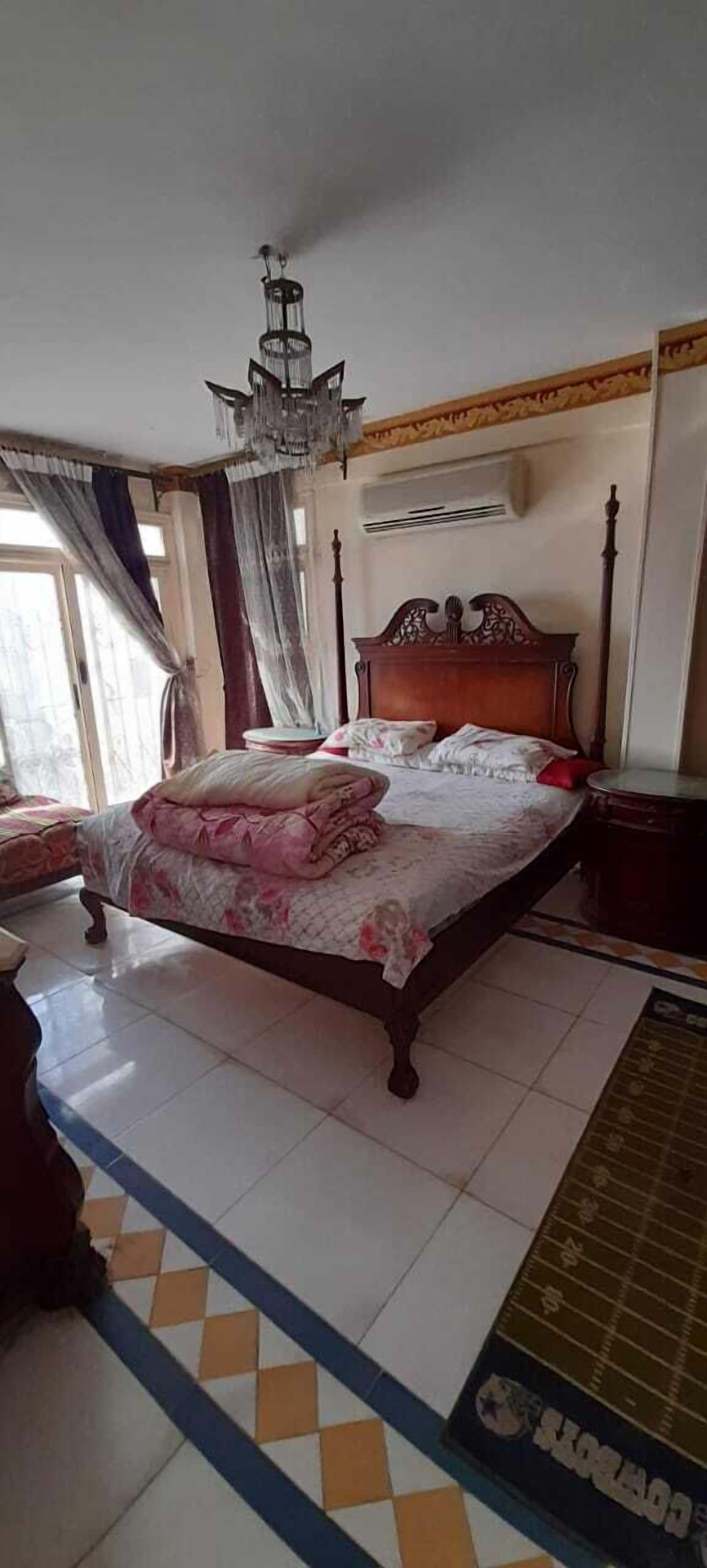 A bedroom with a large bed, a chandelier, curtains, and a tiled floor.