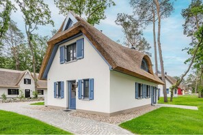 Une maison traditionnelle avec un toit de chaume, des murs blancs et des volets bleus, entourée d’arbres et bordée d’un chemin pavé.