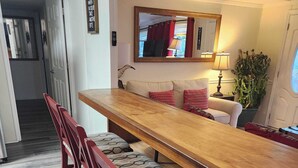 Ein Essbereich mit Holztisch und vier Stühlen, einer Couch mit gemusterten Kissen, einem Spiegel und einer Topfpflanze.