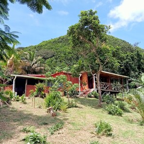 Une maison de campagne aux murs rouges, entourée d’une végétation luxuriante et avec une colline en arrière-plan.