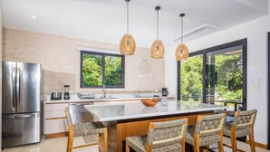 Una cocina moderna con una isla grande, electrodomésticos de acero inoxidable, sillas de mimbre y ventanas amplias con vista a zonas verdes.
