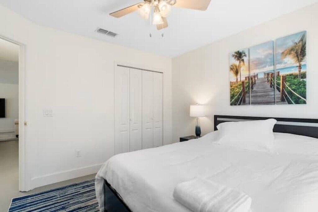 Une chambre d’hôtel avec un lit, un ventilateur de plafond, un tableau représentant une scène de plage et une porte.