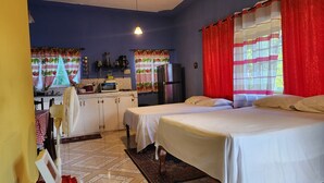 Una habitación compacta con cama, una kitchenette pequeña y ventana con cortinas rojas.