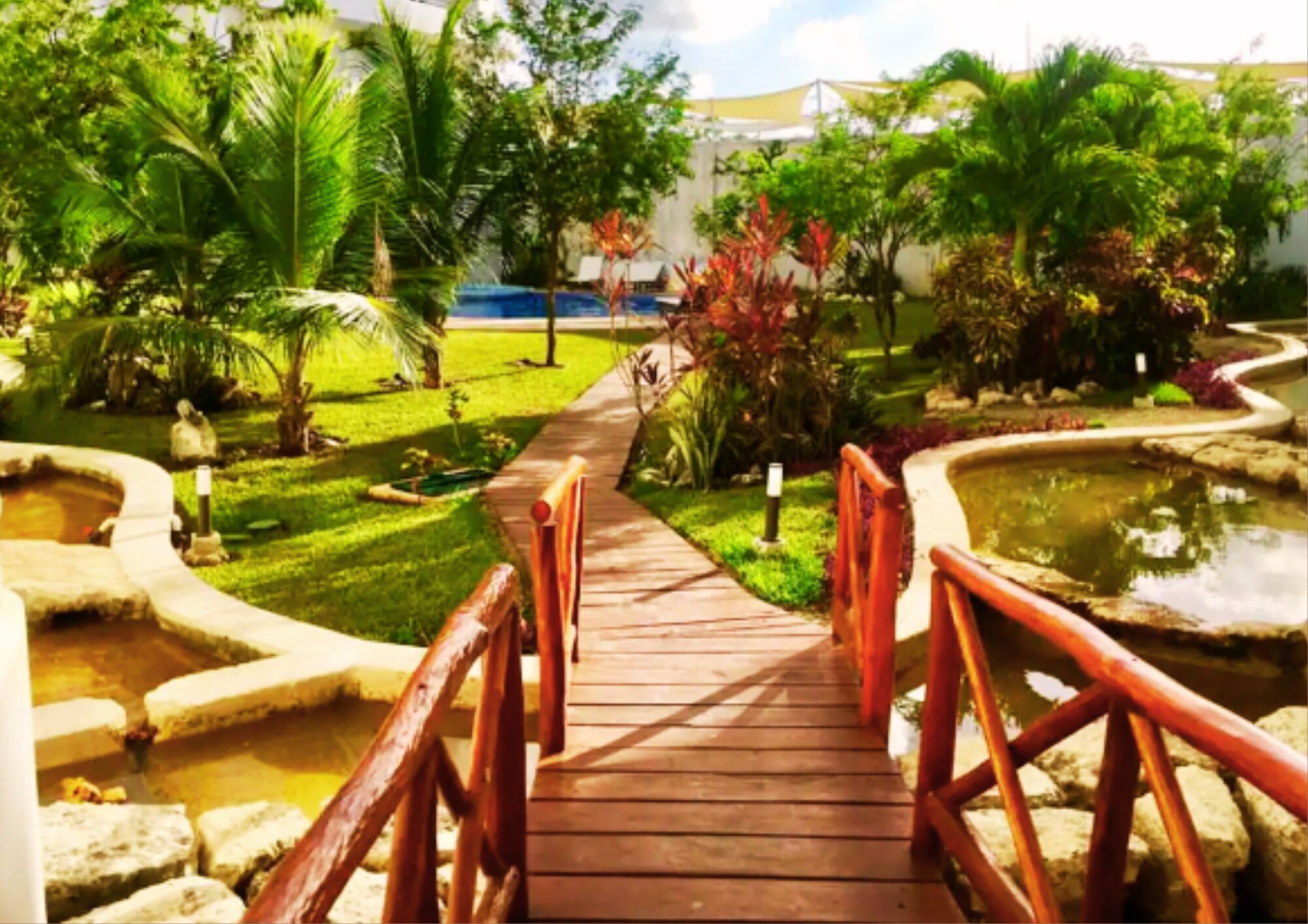Un puente de madera que conduce a una zona de piscina, con un jardín circundante y un edificio al fondo.