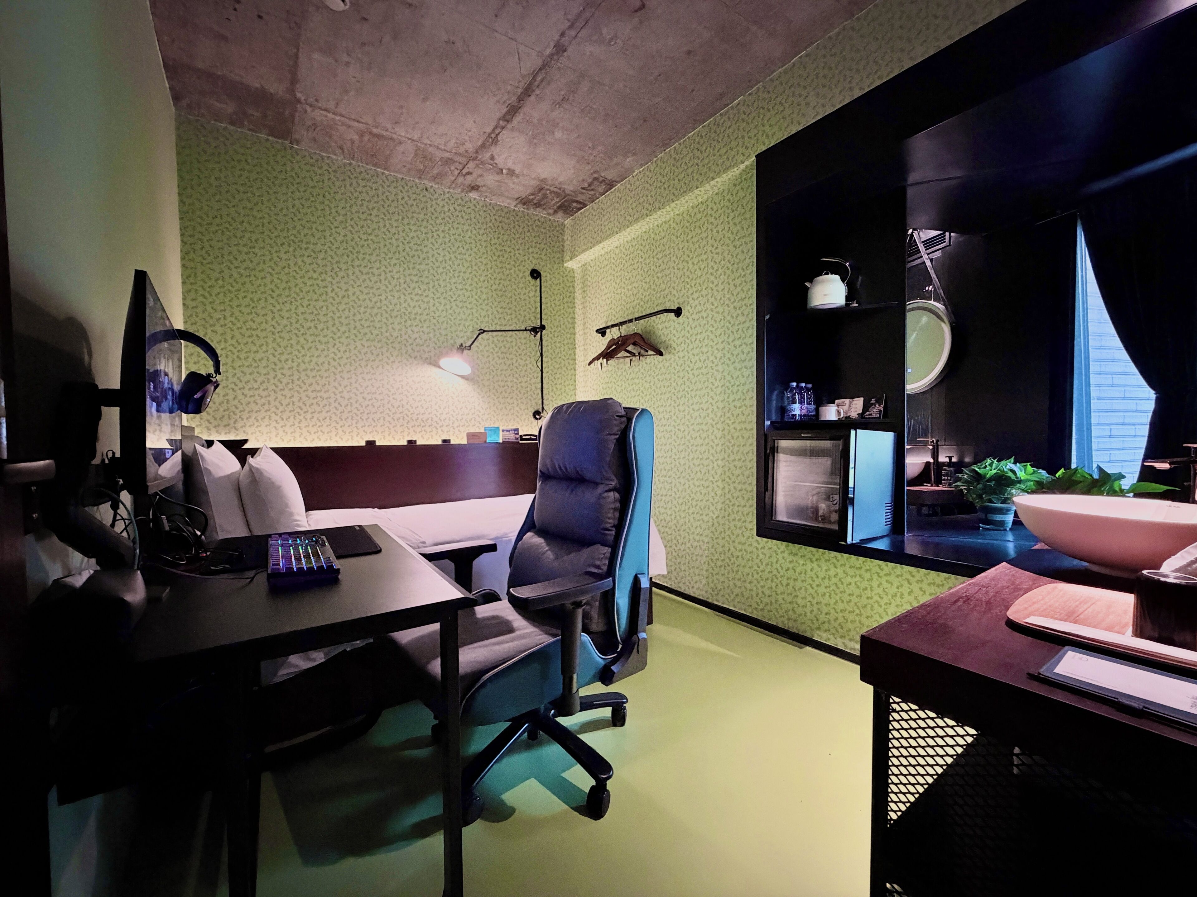 Ein Zimmer mit grüner Wandstruktur, einem Schreibtisch mit Computer und Kopfhörern, einem Stuhl, einem kleinen Kühlschrank und einem Fenster mit Blick.