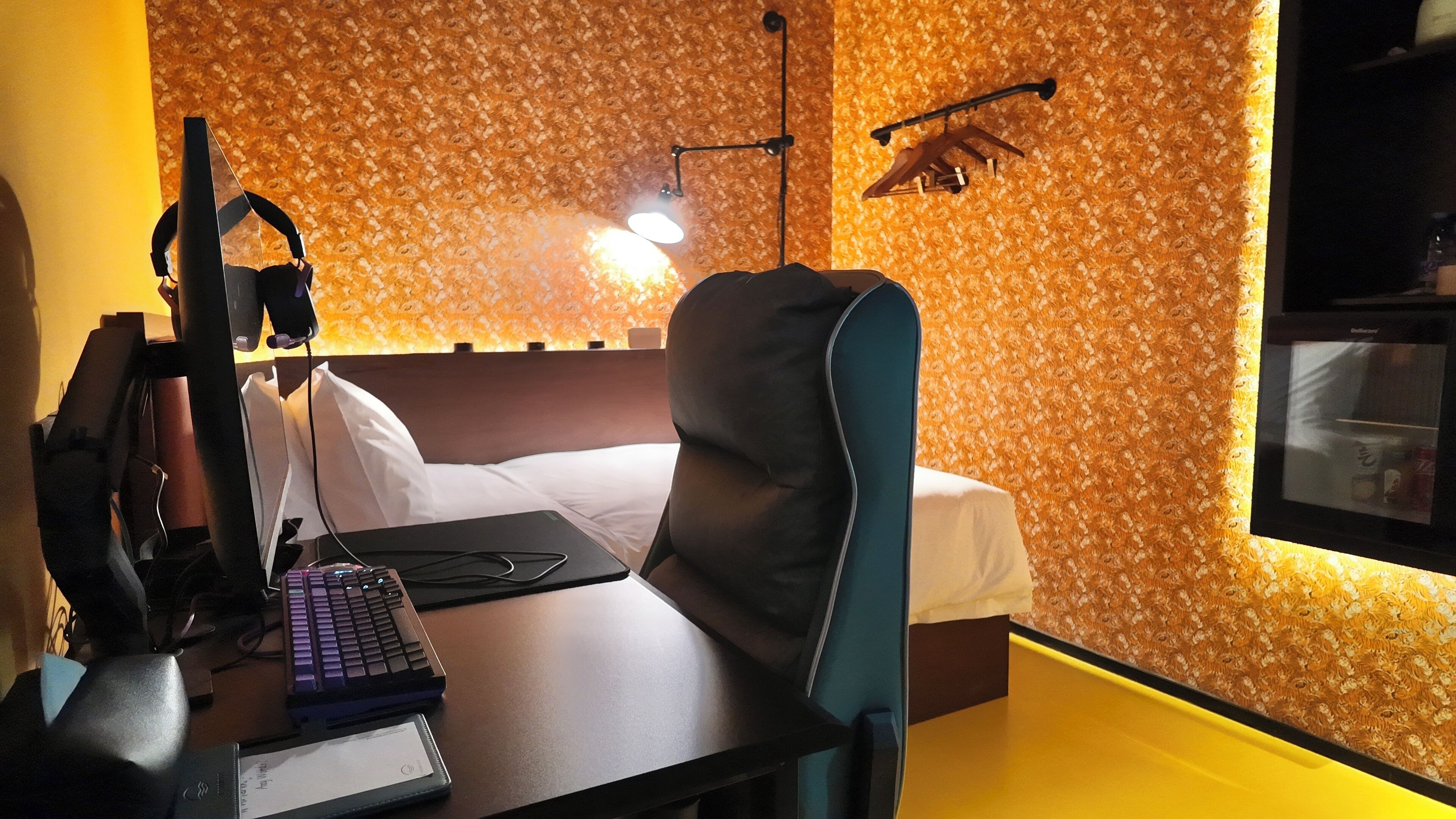 A hotel room with a bed, a desk with a computer and headphones, a lamp, and a window.