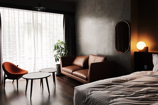 A modern hotel room with a leather sofa, a small round table, a single bed, and a large window with sheer curtains.