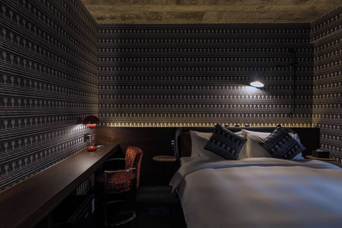 A hotel room with a patterned wall, a bed with pillows, a desk with a lamp, and a chair.