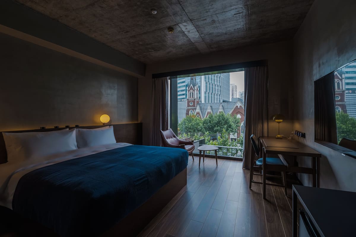 A hotel room with a large bed, a desk, and a chair. There is a view of a cityscape through the window.